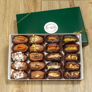 May include: A white box filled with chocolate-covered dates, each topped with nuts and coconut flakes. The box sits beneath a dark green lid with a gold ribbon and a sticker that reads "Box of Dates". The dates are arranged in rows on a wooden surface.