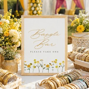 Könnte beinhalten: Ein rechteckiges Schild in einem hellen Holzrahmen mit dem Text Bangle Bar und PLEASE TAKE ONE in goldener und schwarzer Schrift. Das Schild hat eine untere Bordüre aus gelben Wildblumen und ist von Körben mit goldenen Armreifen umgeben.