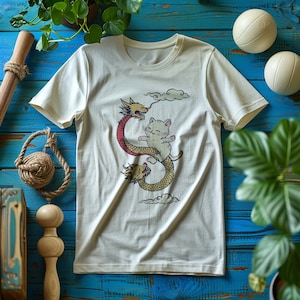 K&ouml;nnte beinhalten: Ein wei&szlig;es T-Shirt mit einem Aufdruck eines Comic-Katzen, das auf einem Drachen reitet. Der Drache ist rot und gr&uuml;n und die Katze ist wei&szlig; mit rosa Ohren und einer rosa Nase. Der Aufdruck ist auf wei&szlig;em Hintergrund.