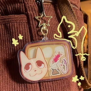 Op de afbeelding: Een heldere acryl sleutelhanger met een kattengezicht en kleine vis- en voedselafbeeldingen erin. De sleutelhanger is bevestigd aan een gouden stervormige sluiting. De achtergrond is een bruine corduroy stof.