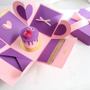 May include: A handmade explosion box in pink and purple, featuring a miniature cake with a pink rose on top. The box has heart-shaped cutouts and decorative elements, including a small gift box with a pink ribbon.
