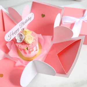 May include: A pink explosion box with a white ribbon and a small pink cake with white and yellow flowers and gold glitter. The box has a heart-shaped opening and the cake has a sign that says "Happy Birthday!"