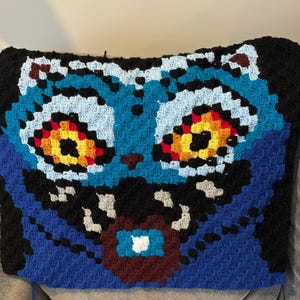 May include: A handmade crochet pillow featuring a stylized cat face. The design uses a pixelated technique with shades of blue, black, white, red, yellow, and brown. The pillow is rectangular and appears to be resting on a gray surface.