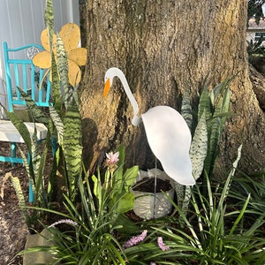 May include: A white metal bird sculpture of a heron with an orange beak, standing in a garden setting with green plants and a white flower.