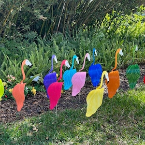 May include: A row of 12 colorful flamingo yard decorations, each with a different color, including yellow, orange, red, pink, blue, purple, and green. The flamingos are standing on a green lawn in front of a lush green background.
