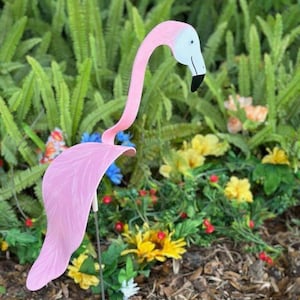 May include: Pink flamingo garden decoration with a white head and black eyes. The flamingo is standing on a metal stake in a garden setting with green plants and flowers.