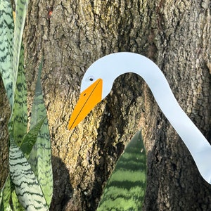 May include: A white paper cutout of a goose head with an orange beak and black eyes. The goose head is partially visible behind a green and white striped plant.