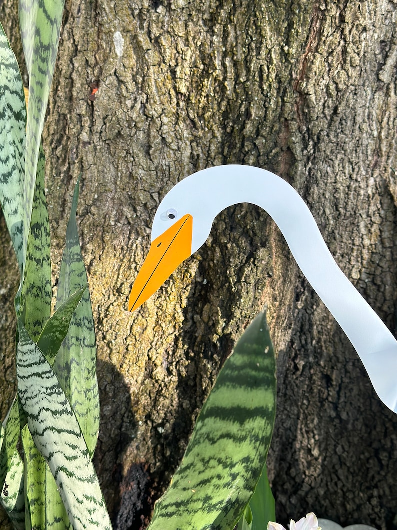 May include: A white paper cutout of a goose head with an orange beak and black eyes. The goose head is partially visible behind a green and white striped plant.
