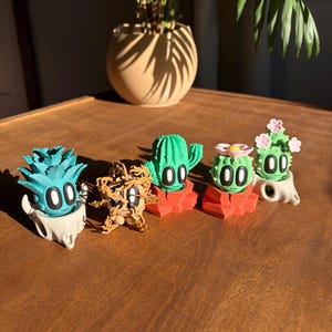 May include: A collection of five whimsical cactus figurines in various colors, including teal, green, and pink. Each figurine has a cartoon-like face with large, oval eyes and is mounted on a small, red or white base. The figurines are arranged on a wooden surface.