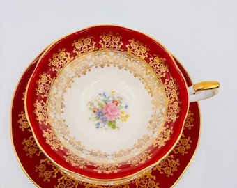 May include: A red and gold teacup and saucer set with a floral design. The cup and saucer have a scalloped edge and a gold trim.