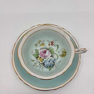 May include: A vintage teacup and saucer set. The cup and saucer are a light teal color with a gold trim. The inside of the cup features a floral design with pink, blue, and yellow flowers. The set is likely made of porcelain.