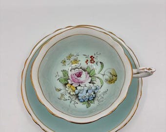 May include: A vintage teacup and saucer set. The cup and saucer are a light teal color with a gold trim. The inside of the cup features a floral design with pink, blue, and yellow flowers. The set is likely made of porcelain.
