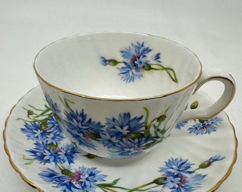 May include: A white teacup and saucer set decorated with a blue floral design. The teacup and saucer have a gold trim. The floral pattern features blue flowers and green stems. The teacup sits on the saucer.