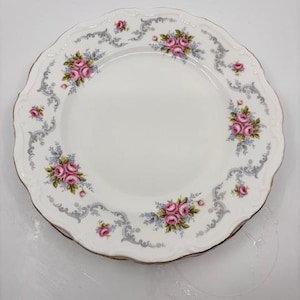 Puede incluir: Plato de porcelana blanca con borde festoneado y borde dorado. El plato está decorado con rosas rosas y detalles azules, sobre un borde gris ornamentado. Un diseño clásico para servir alimentos.