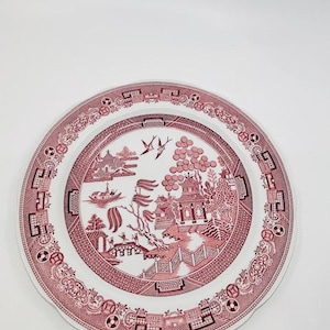 May include: A white ceramic dinner plate with a pink willow pattern. The plate has a scalloped edge and a raised rim.