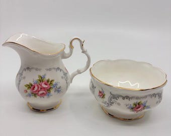 Può includere: Un set di lattiera e zuccheriera in porcellana bianca. Il set presenta un motivo floreale con rose rosa e accenti blu, bordato con finiture dorate. La lattiera ha un manico curvo.