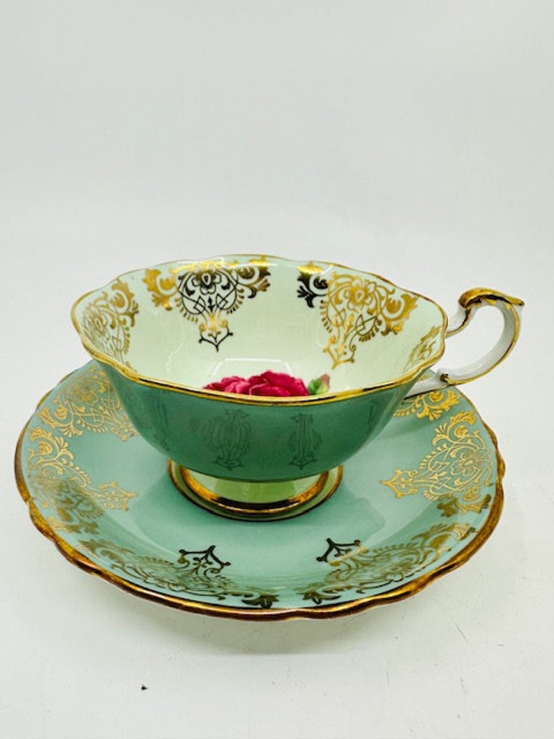 May include: A green teacup and saucer set with gold trim and a pink rose design. The set is made of fine china and has a delicate, elegant look.