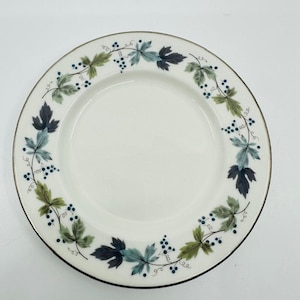 Puede incluir: Un plato de cerámica blanco con un borde decorativo de hojas y bayas verdes y azules. El plato tiene un borde ligeramente elevado.