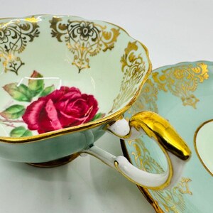 May include: A vintage teacup and saucer set with a delicate floral design. The cup features a pink rose with green leaves and gold accents. The saucer has a matching floral pattern and a light blue background.