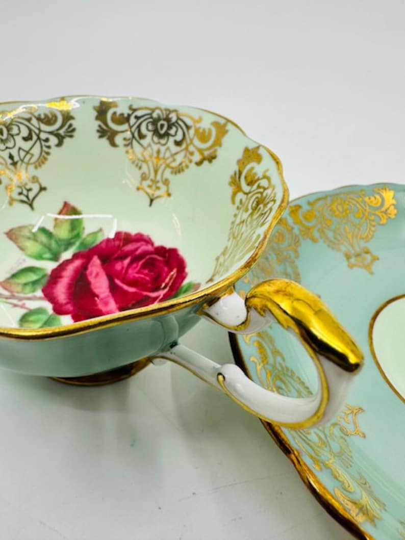 May include: A vintage teacup and saucer set with a delicate floral design. The cup features a pink rose with green leaves and gold accents. The saucer has a matching floral pattern and a light blue background.