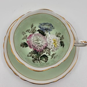 May include: A vintage teacup and saucer set. The cup and saucer are light green with a gold trim. The inside of the cup features a floral design with pink, white, and blue flowers. The handle of the cup is white with gold accents.