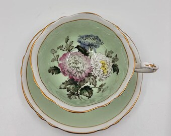 May include: A vintage teacup and saucer set. The cup and saucer are light green with a gold trim. The inside of the cup features a floral design with pink, white, and blue flowers. The handle of the cup is white with gold accents.