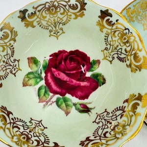 May include: A vintage teacup saucer with a delicate green and gold floral design. The saucer features a single red rose with green leaves and gold accents.