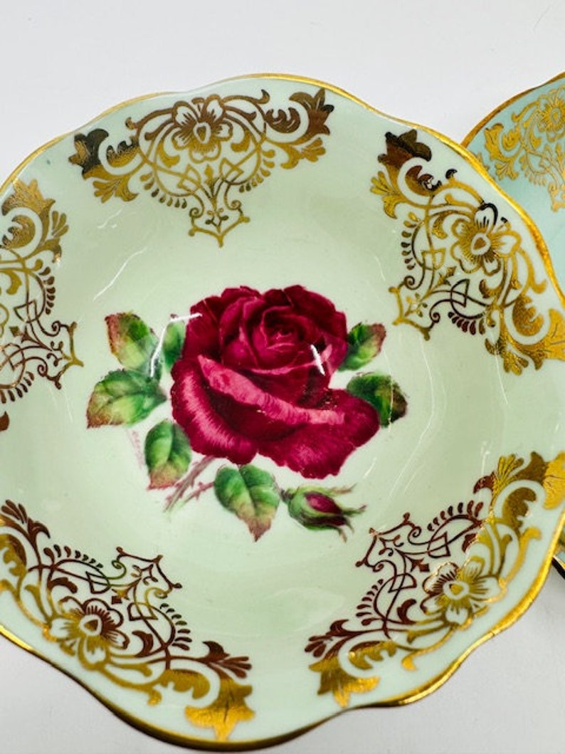 May include: A vintage teacup saucer with a delicate green and gold floral design. The saucer features a single red rose with green leaves and gold accents.