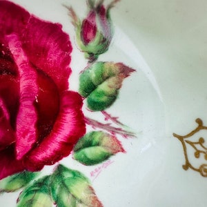 May include: A white porcelain saucer with a hand-painted design of a pink rose with green leaves and a gold trim. The saucer is marked with the text "Remark" in cursive.