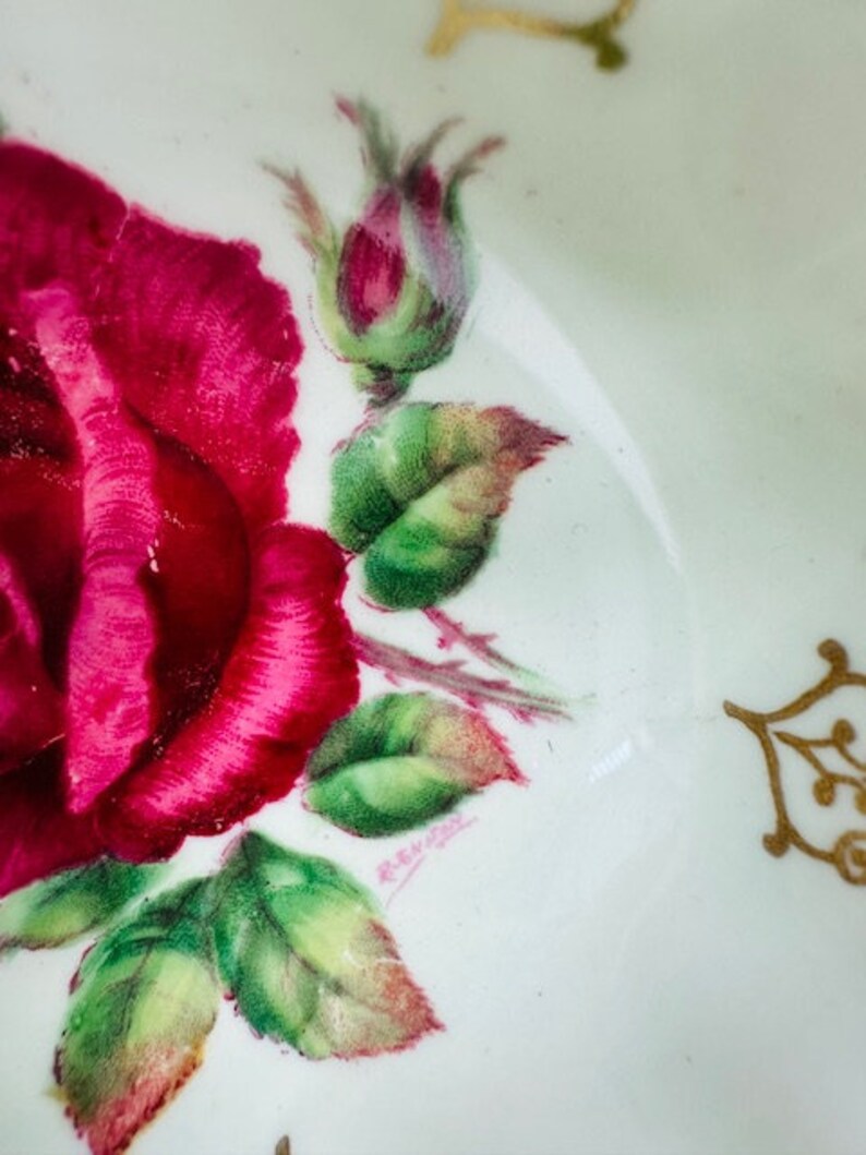 May include: A white porcelain saucer with a hand-painted design of a pink rose with green leaves and a gold trim. The saucer is marked with the text "Remark" in cursive.