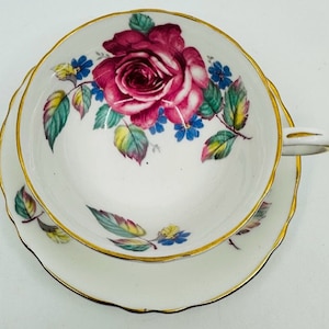 May include: A white teacup and saucer with a gold rim. The teacup features a pink rose with green leaves and blue flowers.