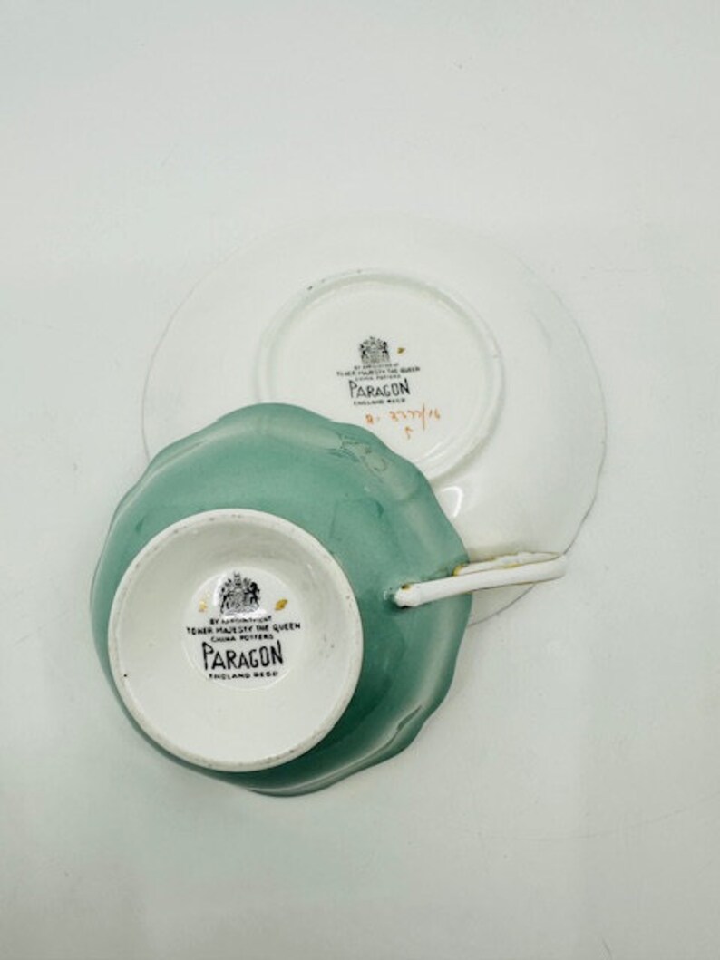 May include: A green teacup and saucer set with a white floral design. The teacup and saucer are made by Paragon, England. The saucer has a gold rim.