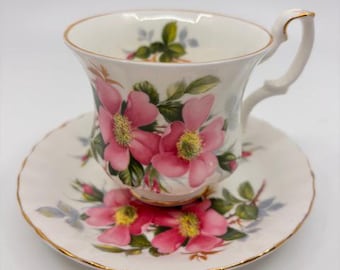 Puede incluir: Juego de taza y platillo de té blanco con diseño floral. La taza y el platillo están decorados con flores rosas, hojas verdes y ribetes dorados. La taza tiene un asa curvada. El platillo es redondo con borde festoneado.