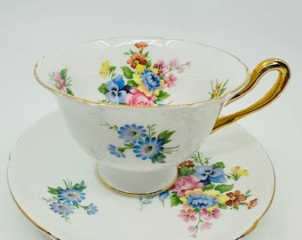 May include: A white teacup and saucer set with a floral pattern. The cup has a gold rim and a gold handle. The saucer has a gold rim and a floral pattern.