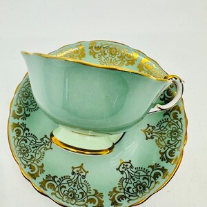 May include: A light green teacup and saucer set with gold floral designs. The teacup has a wide, rounded shape and a delicate handle. The saucer has a scalloped edge and a matching floral design.