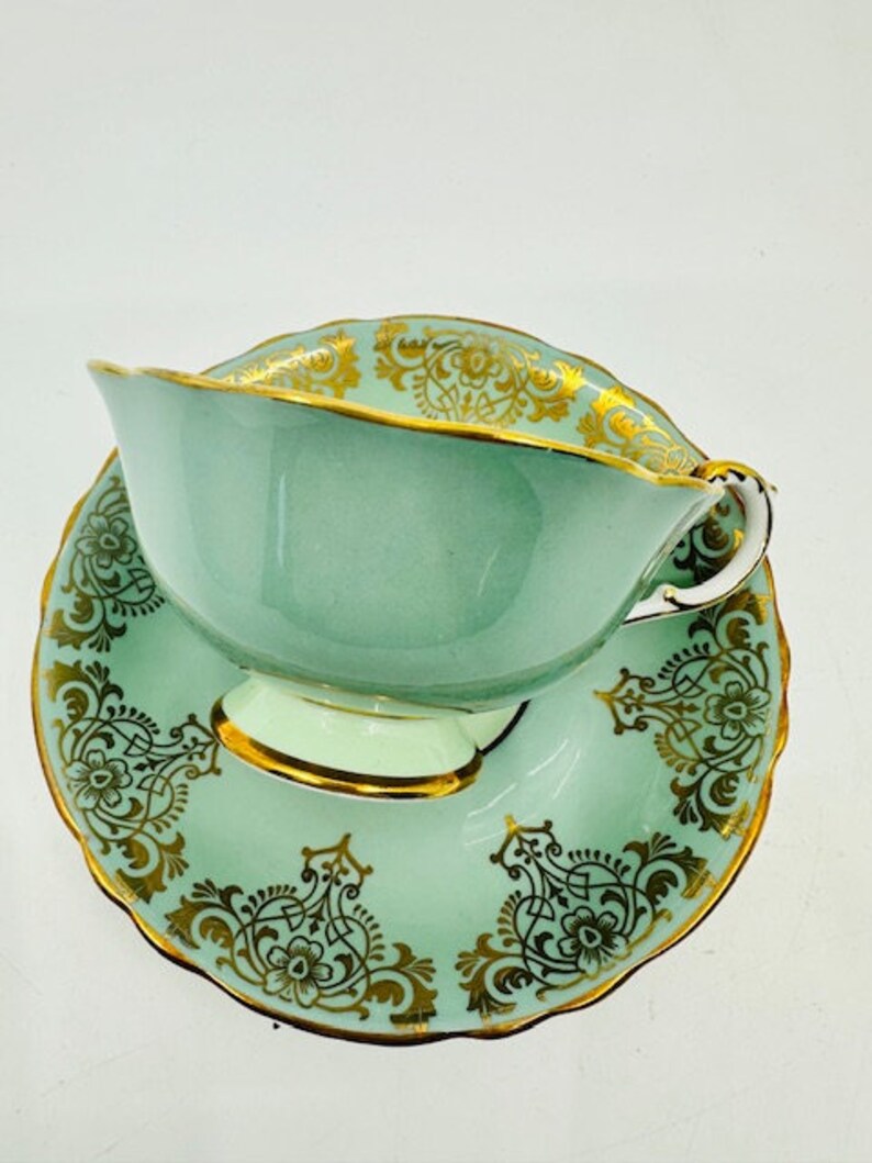 May include: A light green teacup and saucer set with gold floral designs. The teacup has a wide, rounded shape and a delicate handle. The saucer has a scalloped edge and a matching floral design.