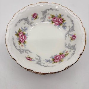 Puede incluir: Plato de porcelana blanca con borde festoneado y ribete dorado. El plato presenta un estampado floral con rosas rosas, detalles azules y volutas grises. Una pieza delicada y elegante para servir o exhibir.