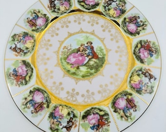 May include: A white porcelain plate with a gold rim and a central design of a couple in a garden setting. The plate is decorated with 12 smaller scenes of couples in various settings around the rim.