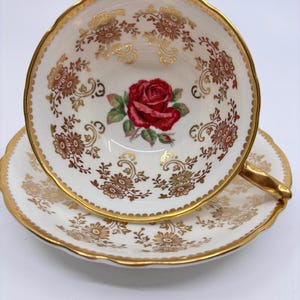 May include: A white teacup and saucer with a gold rim and floral pattern. The cup features a single red rose with green leaves in the center.