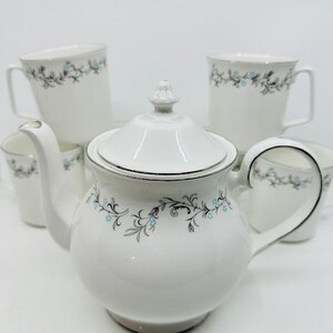 May include: A set of white teacups and teapot featuring a delicate floral design in shades of blue and grey. The teapot has a silver trim around the lid.