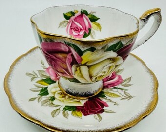 May include: A white teacup and saucer with a gold rim and a floral design featuring pink, white, and red roses. The roses are surrounded by green leaves.
