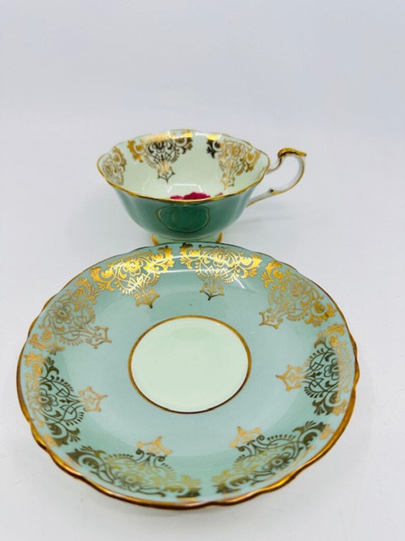 May include: A vintage teacup and saucer set with a delicate floral design in gold and pink on a light green background.