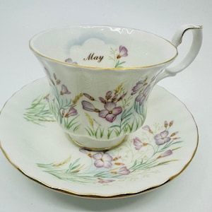 Könnte beinhalten: Eine weiße Teetasse und Untertasse mit einem Blumendesign und dem Wort "Mai" auf der Tasse. Die Blumen sind rosa und weiß und die Blätter sind grün.