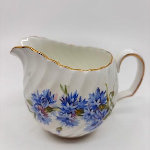 Może przedstawiać: Biały dzbanek do śmietanki z ceramiczną podstawą, złotą obwódką i niebieskim wzorem kwiatowym. Dzbanek ma skręcony kształt i uchwyt.