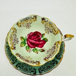 May include: A vintage teacup and saucer set with a delicate floral design. The cup features a single pink rose with green leaves on a light green background. The saucer has a green and gold floral pattern.