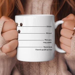 Op de afbeelding: Een witte koffiemok met zwarte tekst die "Shhhh, Almost, Now you may speak, Nevermind, I have to go fill up now" luidt. Boven de tekst staan drie koffieboon-iconen.