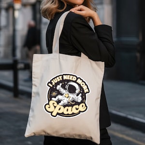 Könnte beinhalten: Eine weiße Canvas-Tragetasche mit einer Schwarzweißillustration eines Astronauten mit dem Text "I just need more space" in einem gelben Kreis.