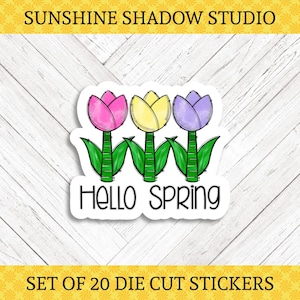 May include: A die-cut sticker featuring three cartoon tulips in pink, yellow, and purple, with green stems and leaves. The text "Hello Spring" is below the flowers. The sticker is on a white background with a wood grain pattern. The text "Sunshine Shadow Studio" is at the top.