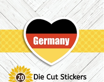 May include: A heart-shaped sticker with the German flag colors: black, red, and yellow. The word "Germany" is in white text. The sticker is on a white wood background with a yellow patterned banner and a sunflower with the number 20 and the words "Die Cut Stickers".