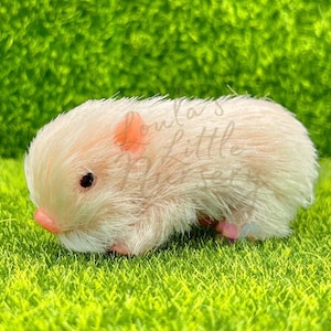 May include: A plush, light pink hamster figurine with a pink nose and ears, and black eyes. The hamster is lying on green artificial grass. The text "Loula's Little Nursery" is visible on the top right of the image.